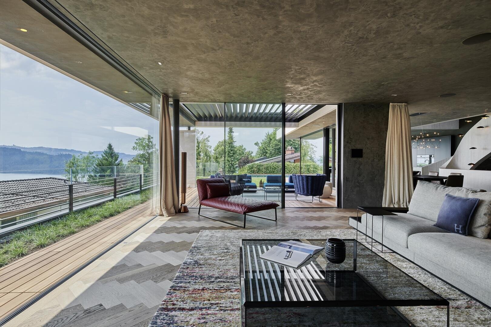 A modern living room with large glass doors that open onto a balcony offering a picturesque view of the hills and trees. The room is furnished with modern furniture, a glass coffee table and a cozy, elegant atmosphere.