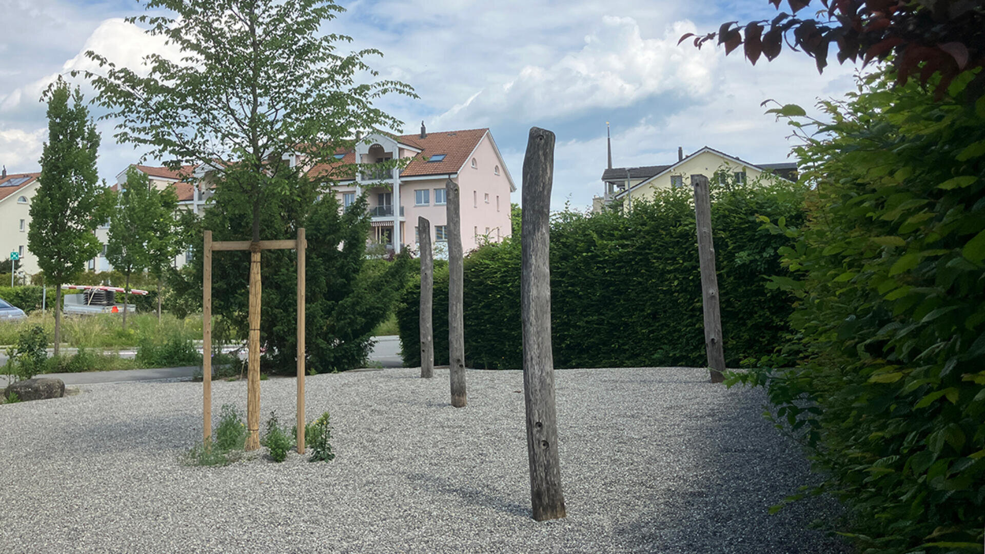Eine kleine Kiesfläche mit mehreren hohen Holzpfosten steht neben einigen jungen Bäumen und Sträuchern, umgeben von grünen Hecken und einigen Wohngebäuden im Hintergrund unter einem teilweise bewölkten Himmel.