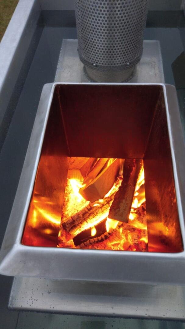 A close-up of burning firewood in a square metal fire pit, in which bright flames and glowing embers can be seen. The fire pit stands on a reflective glass surface outdoors.