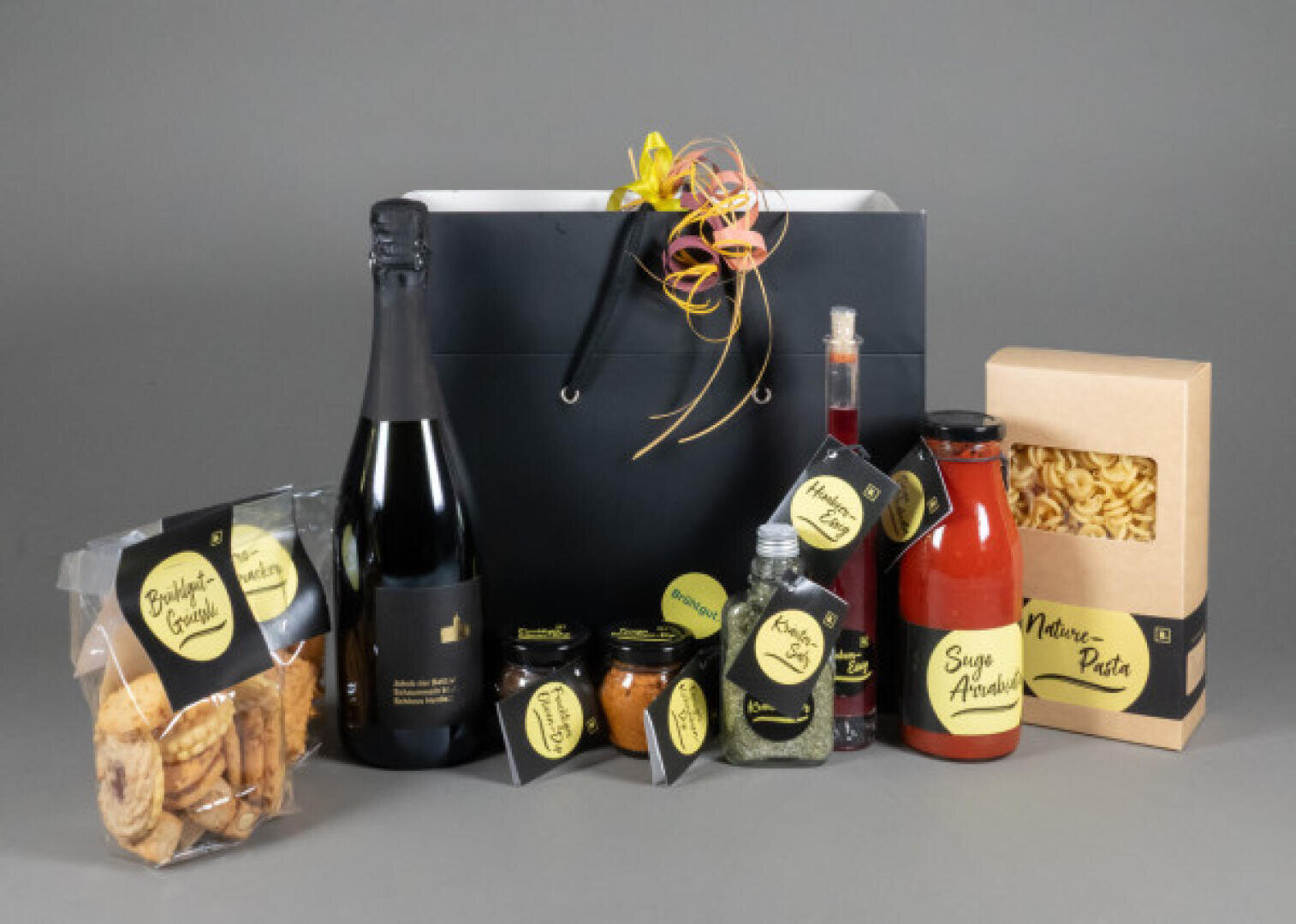 A gourmet gift bag set with a bottle of sparkling wine, glasses of sauces and spices, a box of pasta, dried fruit and bottles of syrup, all neatly arranged against a gray background.