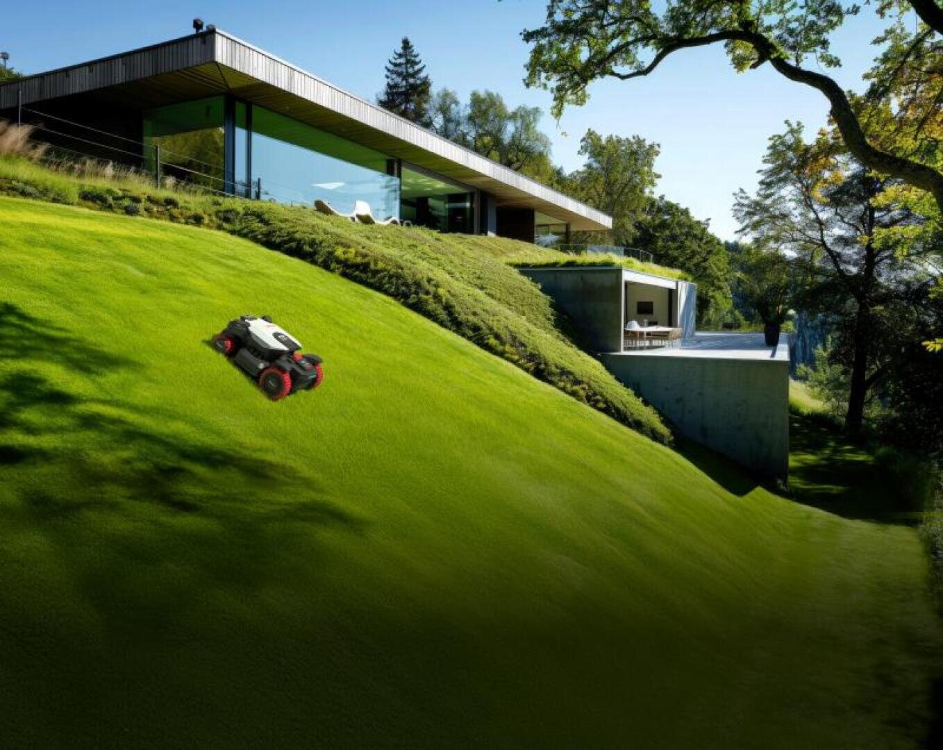 A robotic lawnmower with red wheels works on a steep, green slope in front of a modern house with large glass windows and surrounded by trees.