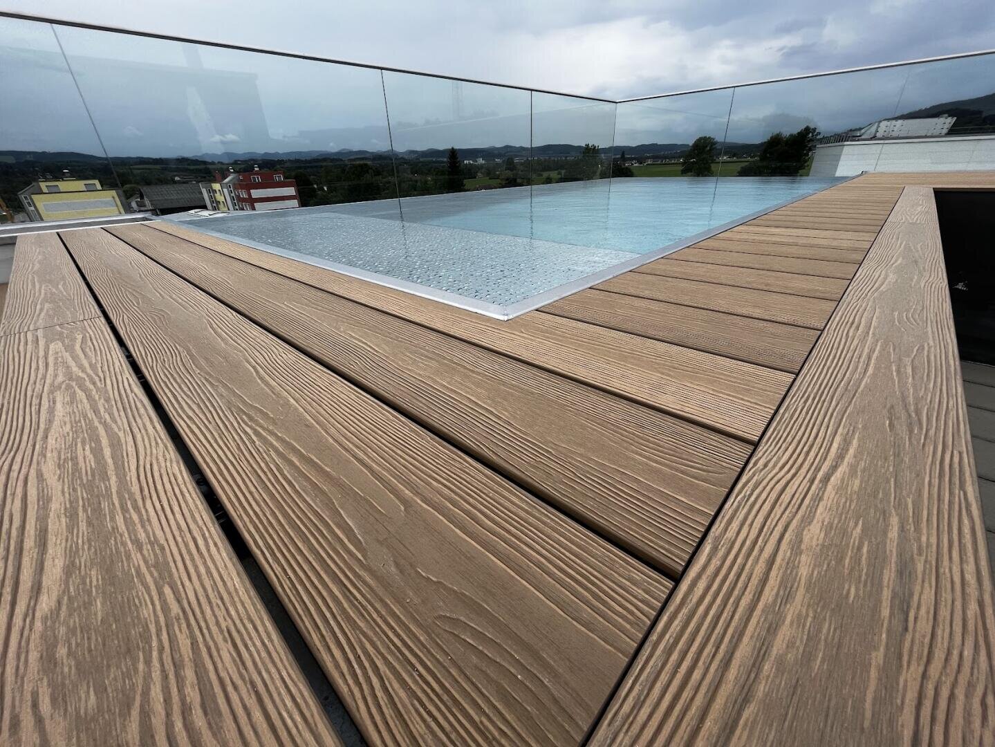 Ein Infinity-Pool auf dem Dach mit einem Geländer aus Klarglas und einer Holzterrasse mit Blick auf Gebäude und Bäume unter einem bewölkten Himmel.