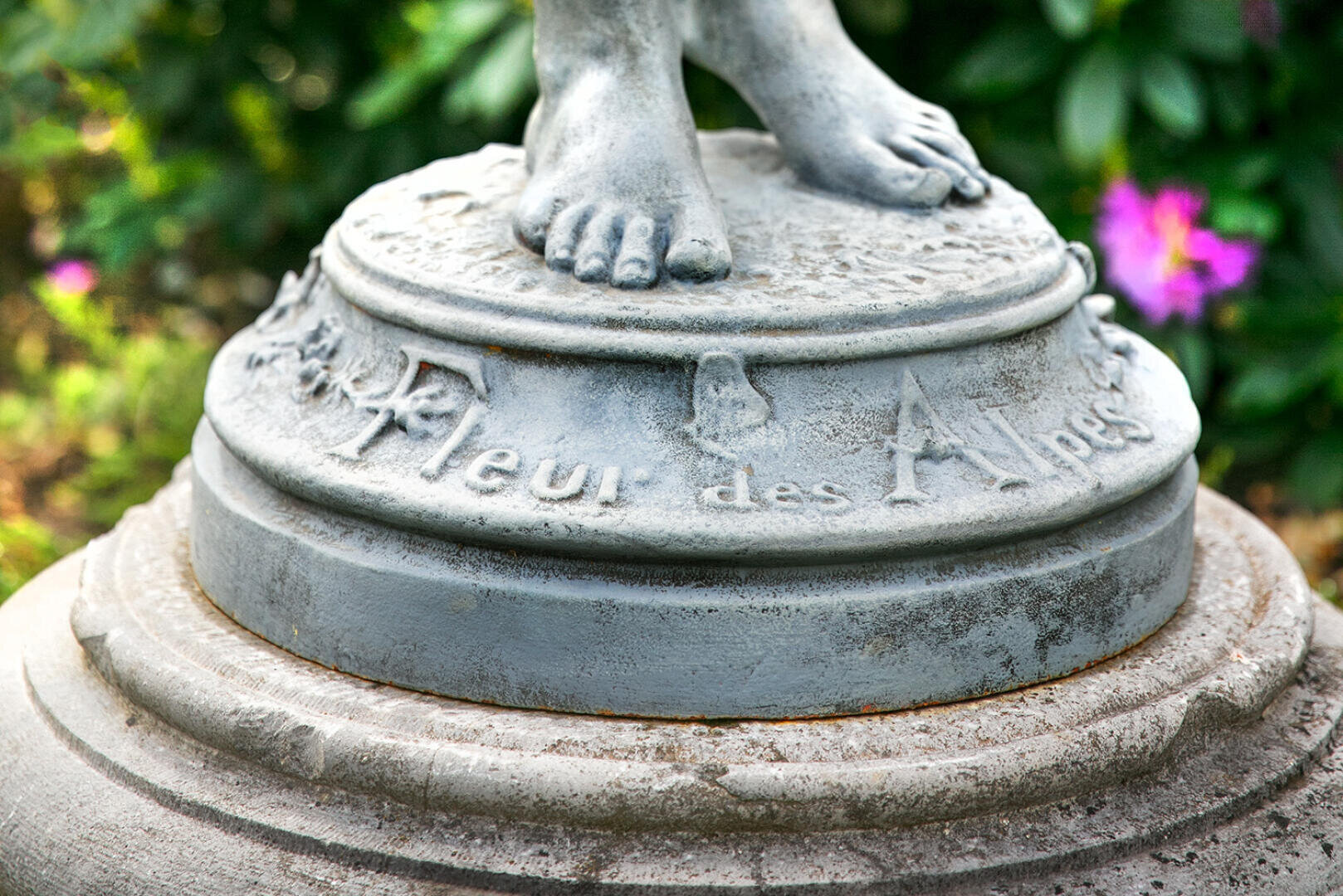 Nahaufnahme des Sockels einer verwitterten Statue mit nackten Füßen, eingraviert mit der Aufschrift Fleur des Alpes und verziert mit geschnitzten Blumenmustern, im Freien mit Grünzeug und einer violetten Blume im Hintergrund.