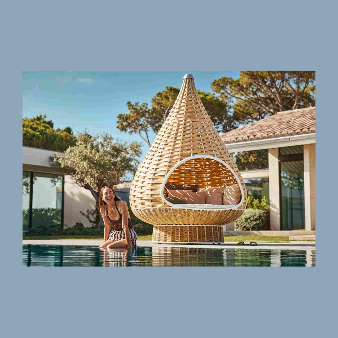 Eine Frau im Badeanzug sitzt lächelnd an einem Pool, hinter ihr ein großes, tropfenförmiges Lounge-Pod aus Weidengeflecht in einer sonnigen Umgebung neben einem modernen Haus.