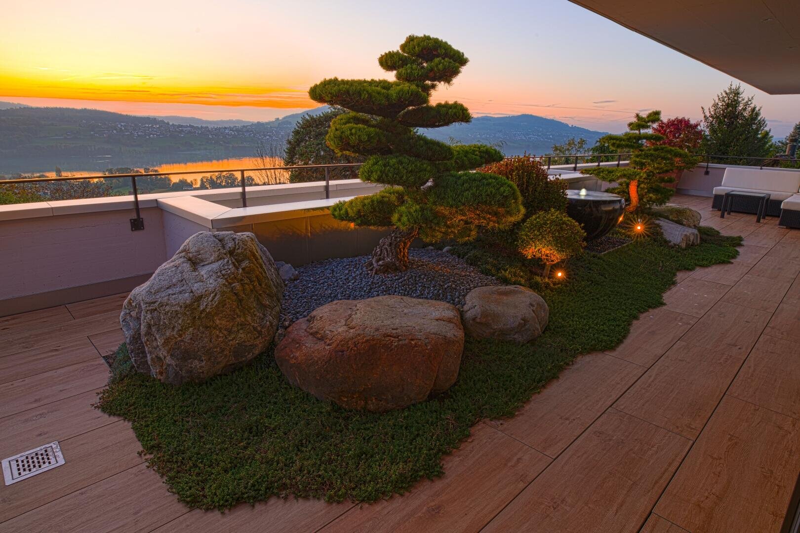Eine landschaftlich gestaltete Terrasse mit gepflegten Sträuchern, großen Steinen und stimmungsvoller Beleuchtung bietet einen Blick auf einen malerischen See und die Hügel bei Sonnenuntergang mit warmen orangefarbenen und violetten Tönen am Himmel.