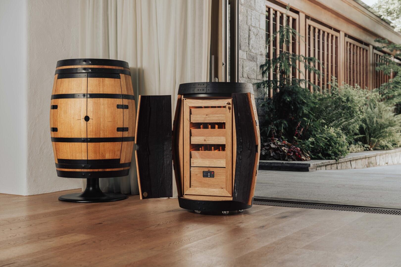 Two wooden barrels stand inside; one is closed and standing upright, the other is open and provides a view of shelves with wine bottles. Large windows and lush green areas can be seen in the background.