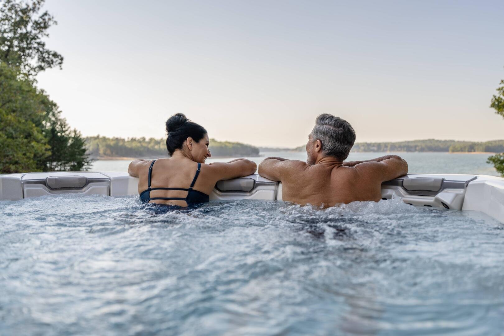 Eine Frau und ein Mann entspannen sich in einem Whirlpool im Freien, schauen sich an und lehnen sich an den Rand. Der Whirlpool überblickt einen malerischen, von Bäumen umgebenen See unter einem klaren Himmel.