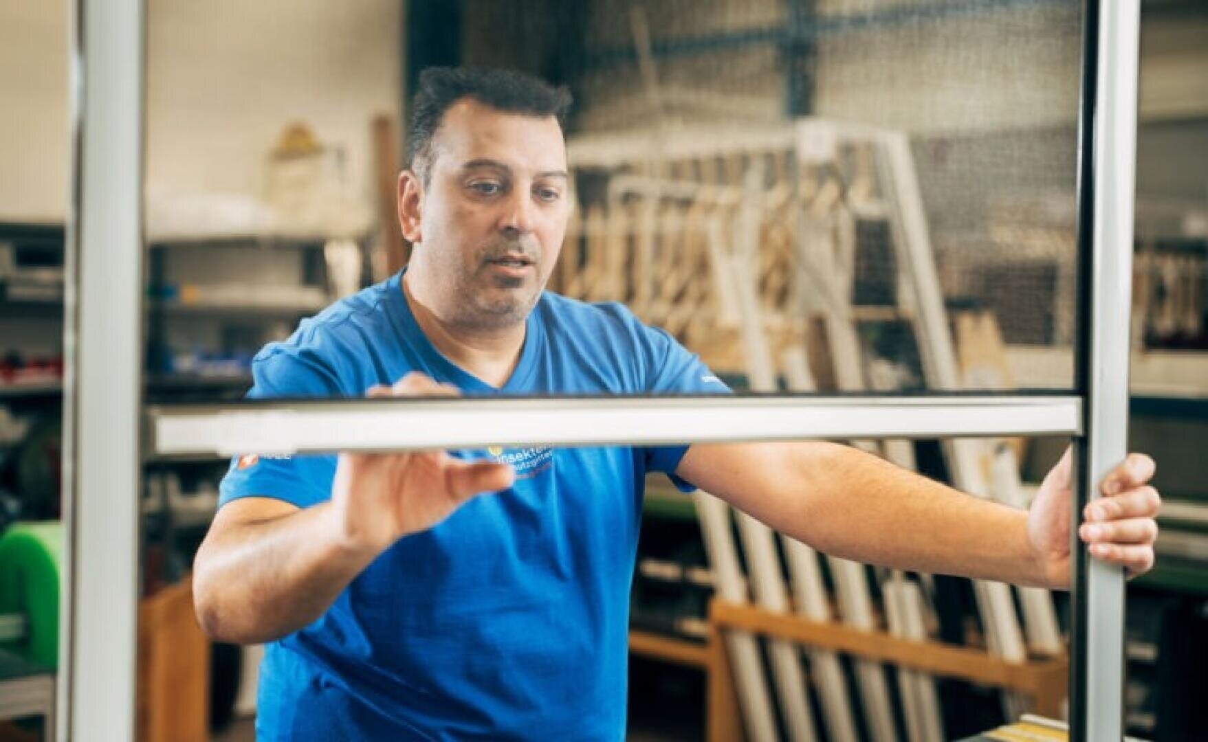 Ein Mann in einem blauen Hemd montiert oder prüft einen Fensterrahmen in einer Werkstatt mit Werkzeugen und Geräten im Hintergrund.