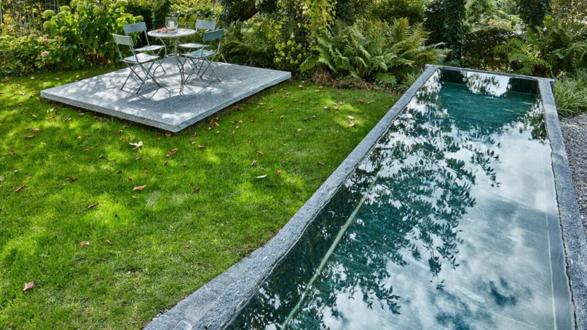 A small reflecting pool with clear water sits on a grassy lawn beside a stone patio with a metal bistro table and two chairs, surrounded by lush greenery and garden plants.