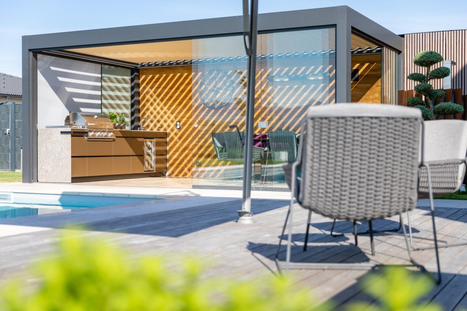 Moderne Außenküche mit Grill, Esstisch und Stühlen unter einer eleganten Pergola. Das Sonnenlicht wirft gestreifte Schatten auf die Holzwand. Im Vordergrund sind Terrassenmöbel und ein Pool zu sehen.