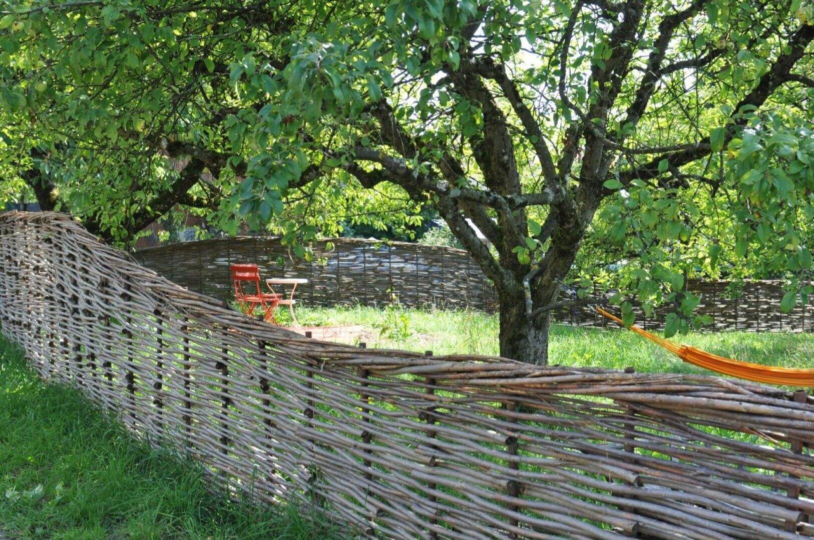A rustic woven wooden fence winds its way through a grassy garden with deciduous trees. Inside the fence are a red chair and a small table, and part of an orange hammock can be seen on the right-hand side. The sunlight filters through the foliage.