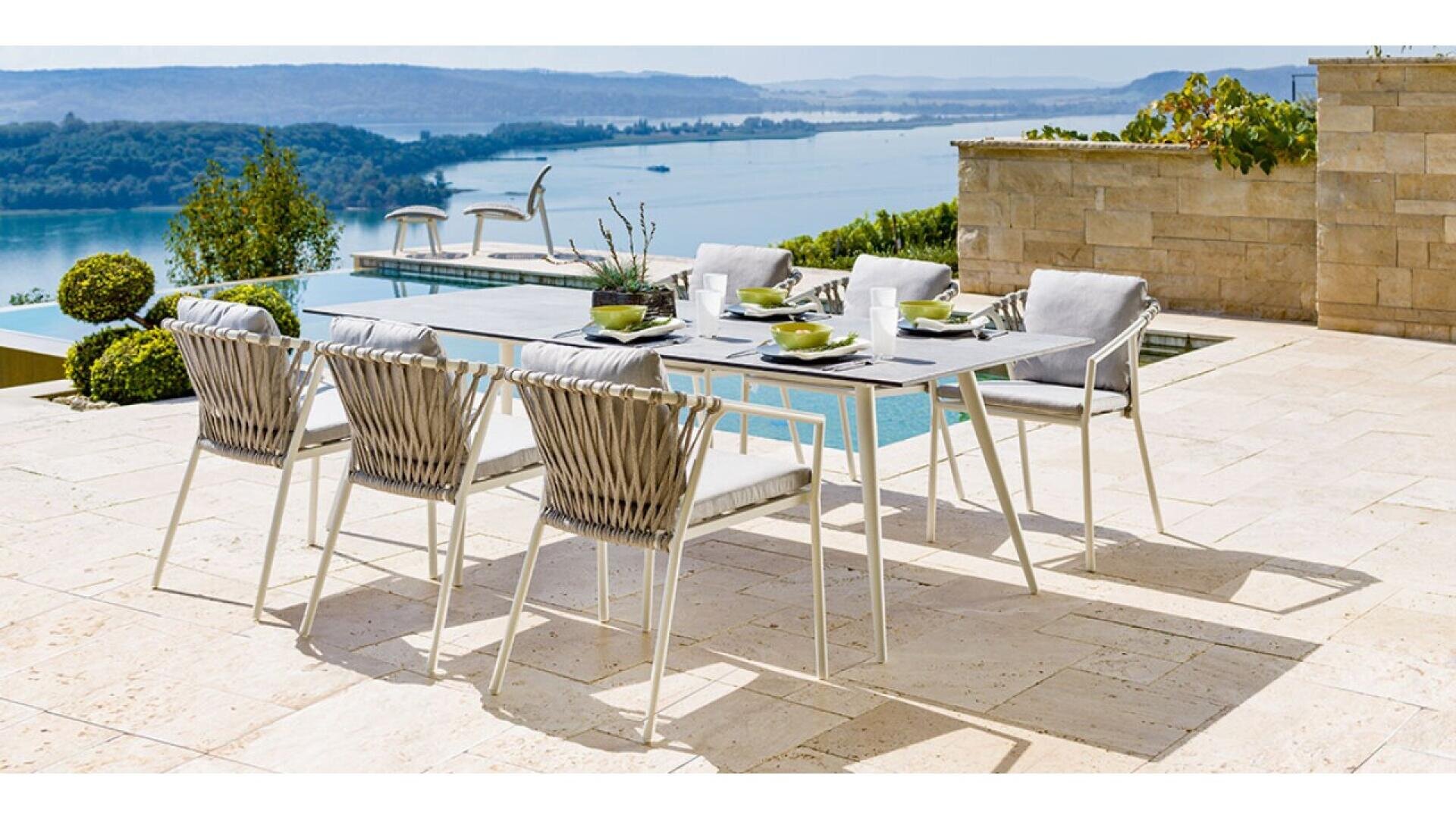 Outdoor dining table with six cushioned chairs set beside a pool, overlooking a scenic view of a lake and distant hills under a clear sky. Table is set with plates, cups, and a centerpiece.