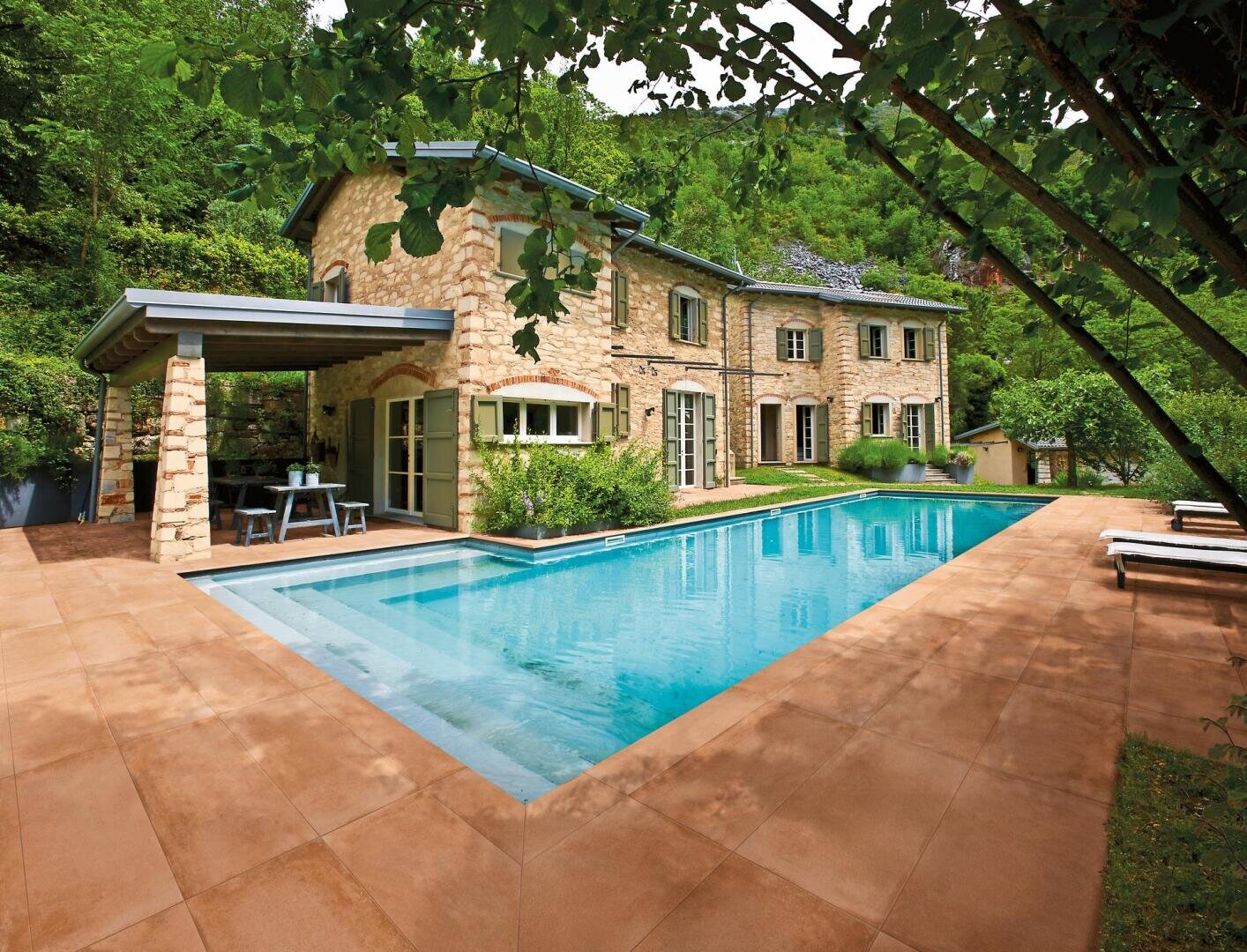 Ein Steinhaus mit großen Fenstern und einer überdachten Terrasse liegt neben einem rechteckigen Swimmingpool, umgeben von Bäumen und Liegestühlen auf einer gefliesten Terrasse in einer üppigen, grünen Umgebung.