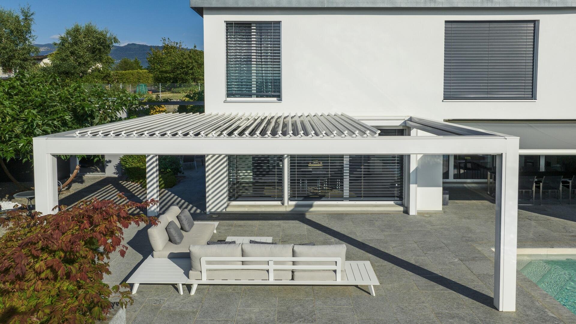 A modern patio with a white pergola, outdoor sofa, coffee table, and chairs on a stone-tiled terrace next to a pool, attached to a white house with large windows and closed blinds.
