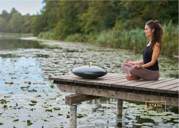 Eine Frau sitzt im Schneidersitz auf einem hölzernen Steg über einem mit Seerosenblättern bedeckten See und meditiert, während neben ihr eine moderne schwarze Feuerschale brennt. Im Hintergrund stehen üppig grüne Bäume. Der Text STAHL BLAU steht in der unteren rechten Ecke.