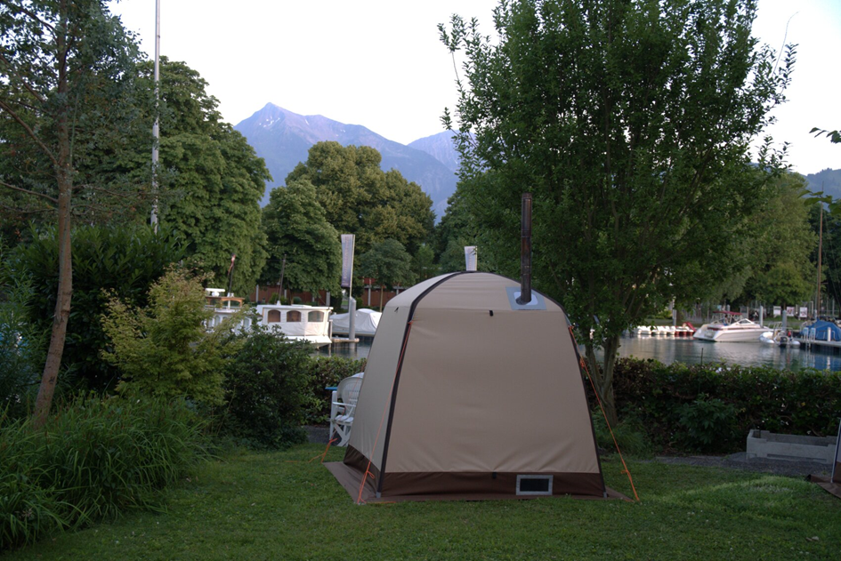A beige tent stands on a grassy area near a body of water, surrounded by trees and greenery. Boats are docked along the water, and mountains are visible in the background under a clear sky.