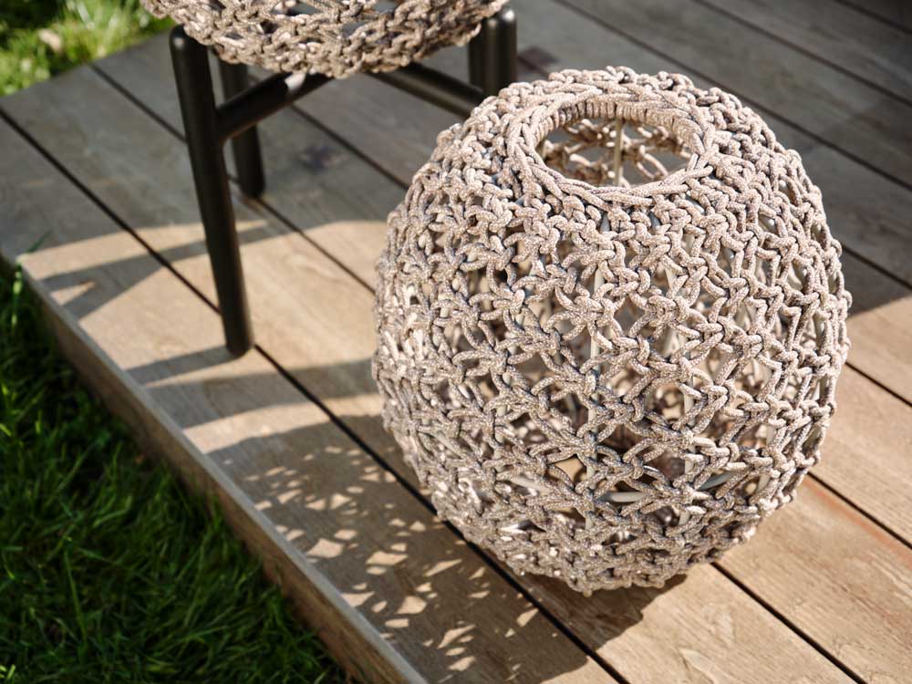 Eine runde, geflochtene Laterne mit einem komplizierten, geknüpften Muster steht auf einem Holzdeck neben einem Hocker. Das Sonnenlicht wirft gemusterte Schatten auf die Terrasse, und das nahe gelegene Gras ist teilweise sichtbar.