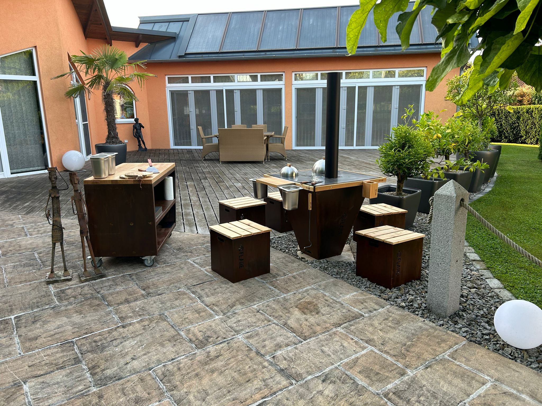 Moderner Innenhof mit Holz- und Metallm&ouml;beln, darunter ein Ofen, Hocker und ein Rollwagen. Eine getopfte Palme, Gr&uuml;npflanzen und eine gro&szlig;e Statue schm&uuml;cken den Raum, mit einem Haus und gro&szlig;en Fenstern im Hintergrund.