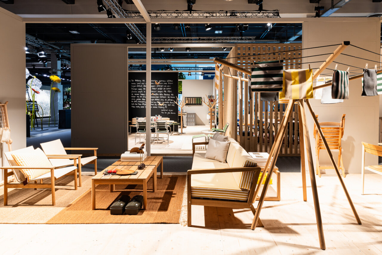 In einem modernen Ausstellungsraum werden Holzmöbel ausgestellt, darunter Sofas, Stühle, Tische und ein Wäscheständer mit gestreiften Textilien. Warme Beleuchtung und neutrale Töne schaffen eine gemütliche, einladende Atmosphäre.