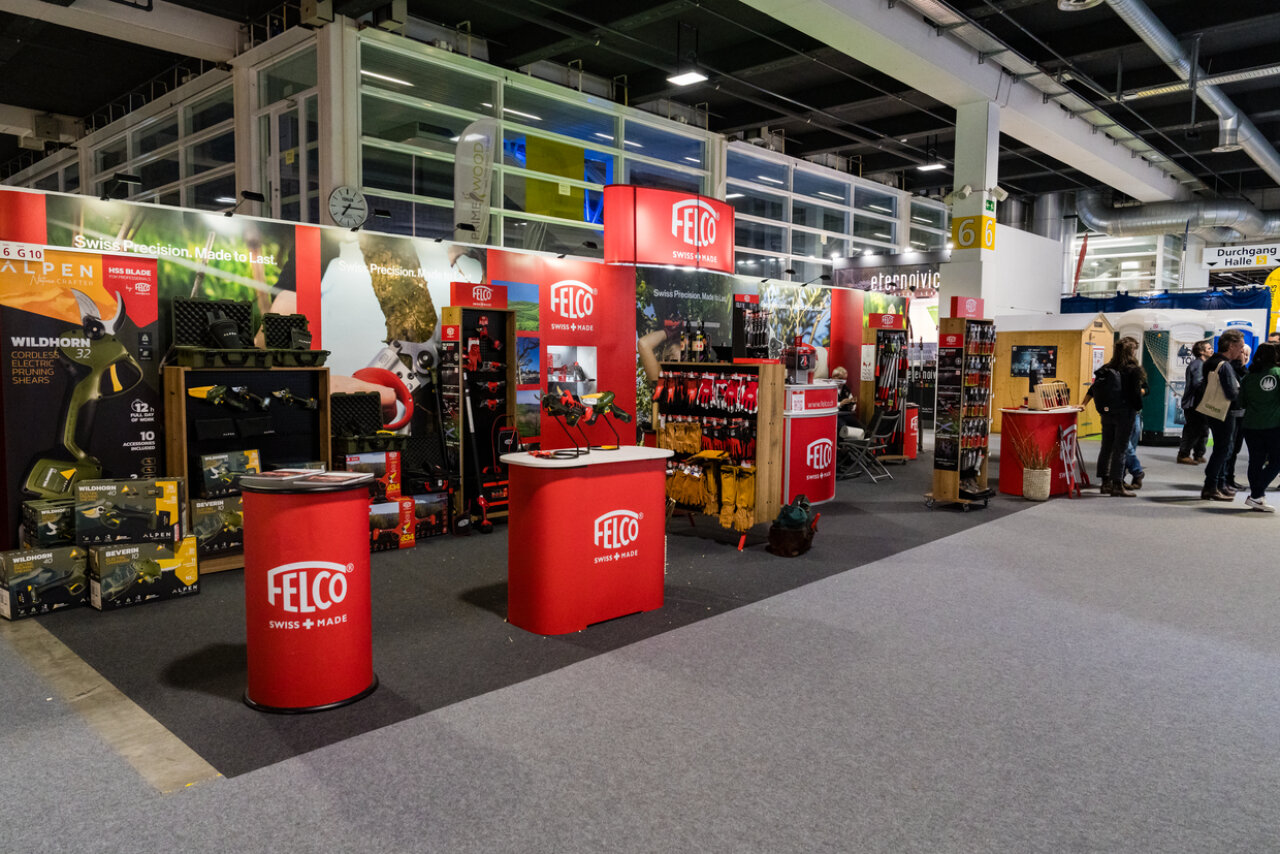 Auf einem Messestand zeigt FELCO rote Displays mit Werkzeugen, Bannern und Werbematerialien. An den Ständen werden Produkte ausgestellt, und die Besucher erkunden die Ausstellungshalle, während im Hintergrund verschiedene andere Stände zu sehen sind.