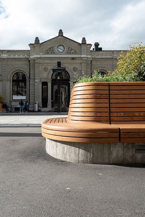 A modern curved wooden bench with plants sits in front of a historic stone building with a clock above the entrance under a cloudy sky.