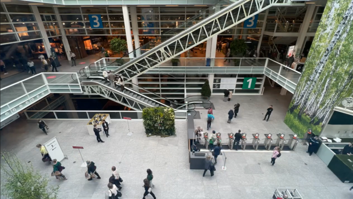 A spacious, modern building interior with escalators, glass walls, numbered signs, and people walking or waiting near a reception area. Large windows reveal indoor plants and upper floors.