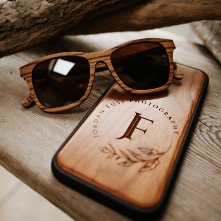 Eine hölzerne Sonnenbrille und ein hölzernes Telefonetui mit der Gravur von Jordan Fuss Photography und einem dekorativen F ruhen auf einer rustikalen Holzoberfläche.