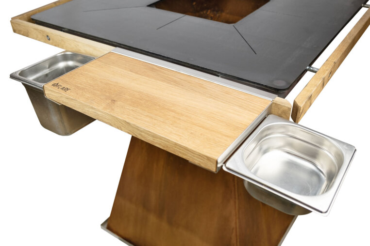 Close-up of a wooden and metal barbecue table for outdoor use with two stainless steel trays on either side and a flat metal cooking surface on top.