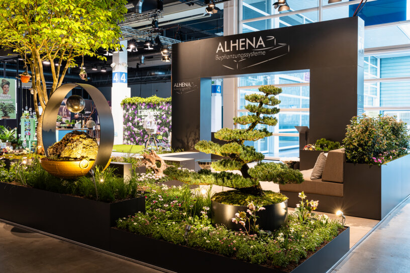 Eine moderne Indoor-Gartenausstellung mit gepflegten Bäumen, üppigem Grün, einer moosbewachsenen Kugel in einem Metallring, Bänken und einem großen schwarzen Ausstellungsrahmen mit dem Schriftzug ALHENA Begrünungssysteme über dem Kopf.