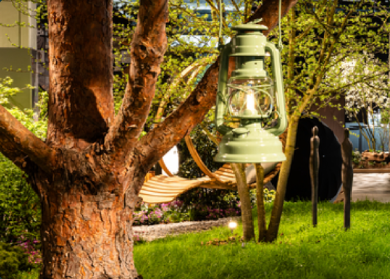 Eine grüne Laterne hängt an einem Baum in einem üppigen Garten mit Gras, Pflanzen und einer Holzschaukel im Hintergrund. Das warme Licht der Laterne schafft eine gemütliche Atmosphäre.