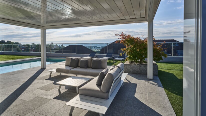 Modern outdoor patio with a shaded seating area featuring cushioned sofas and a coffee table, next to a pool; glass railing and rooftops with a scenic view in the background under a partly cloudy sky.