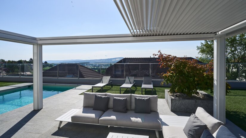 Modern outdoor patio with white sofas and gray cushions, next to a swimming pool. There are lounge chairs facing a scenic city view, and a large planter with a small tree. The area is shaded by a slatted pergola.