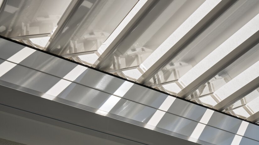 Sunlight streaming through white angled slats of a modern louvered roof, casting geometric shadows on the surfaces below.