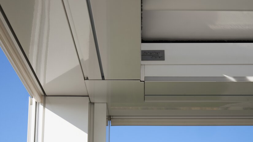 Close-up view of a modern, white metal architectural structure with clean lines and glass panels, set against a clear blue sky. A small metal plaque is attached to the frame.