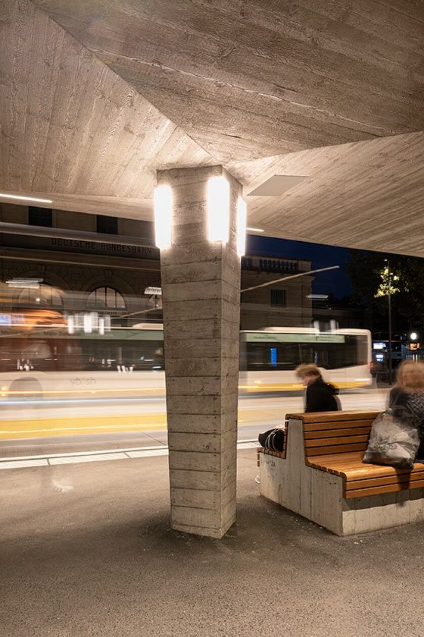 A concrete bus stop with wooden benches at night. Two people are sitting on the benches, and a bus speeds by, appearing blurred. Bright lights illuminate the area, and a building is visible in the background.