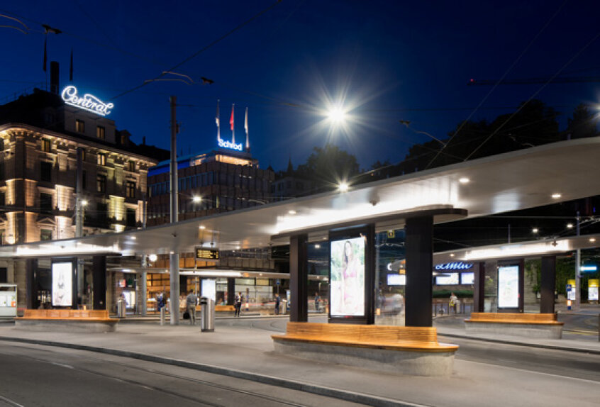 A modern tram station at night with illuminated benches and advertisement boards, set against historic buildings with glowing signs. The sky is dark blue and streetlights brighten the scene.