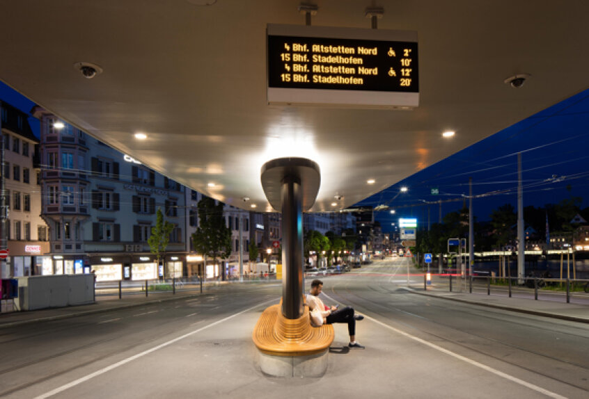 A person sits alone on a modern wooden bench under a large canopy at a tram stop in a city at dusk, with an electronic schedule board displaying tram times overhead. The street around is mostly empty and illuminated.