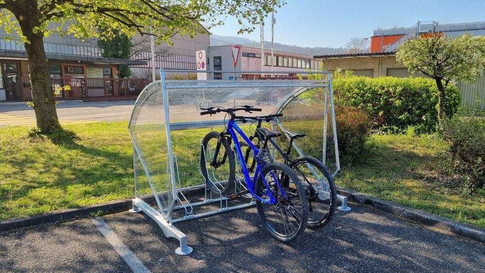 Zwei Fahrräder sind unter einem transparenten Metallunterstand auf einem Parkplatz geparkt, mit grünem Gras, Bäumen und Industriegebäuden im Hintergrund an einem sonnigen Tag.