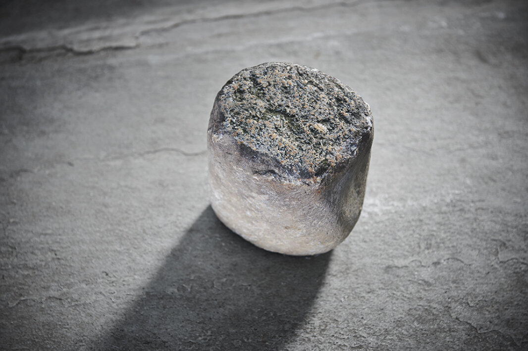A single rough, cylindrical stone with a flat top lies on a textured gray surface and casts a soft shadow to the left. The stone has a blotchy, weathered appearance.