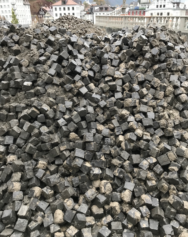 A large pile of small, dark, square paving stones, mixed with dirt, stacked in an urban outdoor area with visible buildings in the background.