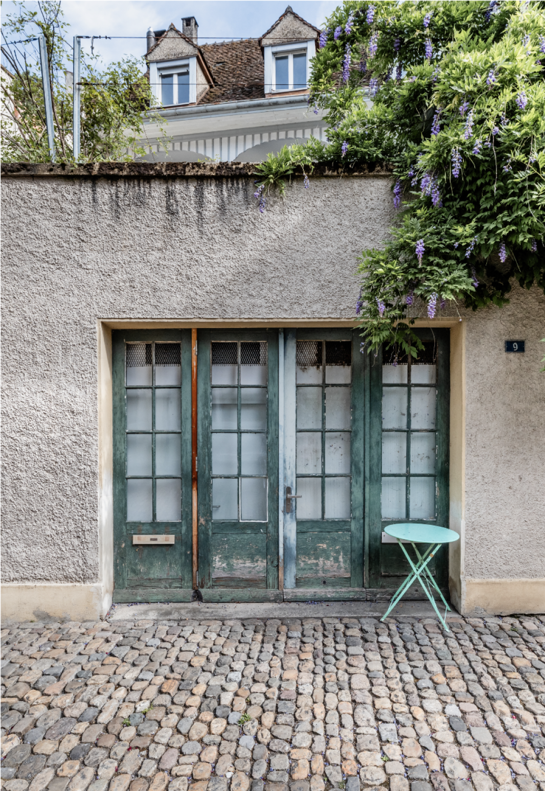 A pair of weathered green double doors in a textured gray wall, with a small round mint green table on the right. Purple wisteria flowers hang above, the floor is covered with cobblestones. A house with dormer windows can be seen behind the wall.