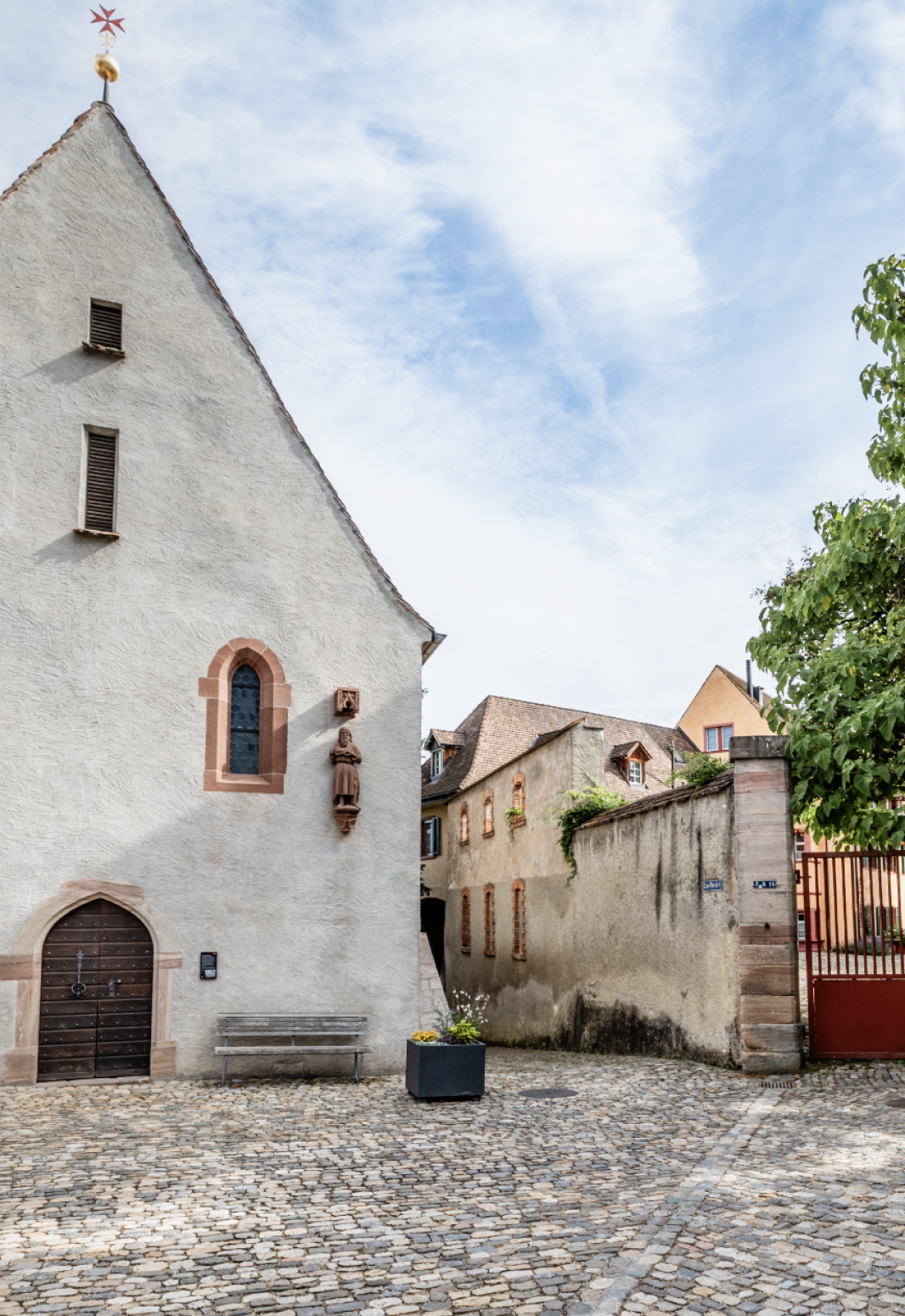 A cobblestone courtyard with a white church with arched windows and a wooden door, a bench, a flower pot and adjoining old stone buildings under a partly cloudy sky.