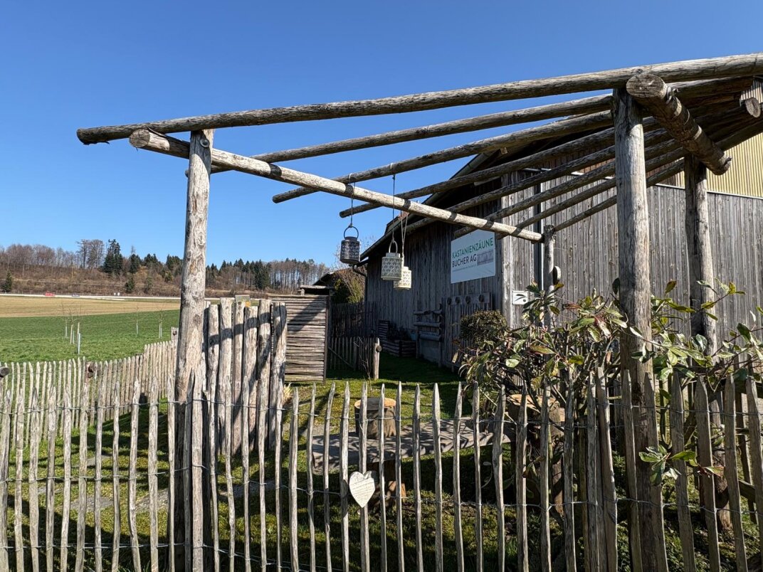 Eine rustikale Holzkonstruktion mit einer Pergola und einem Lattenzaun steht neben einer Wiese unter blauem Himmel; Topfpflanzen und hängende Dekorationen sind in der Nähe des Eingangs zu sehen.