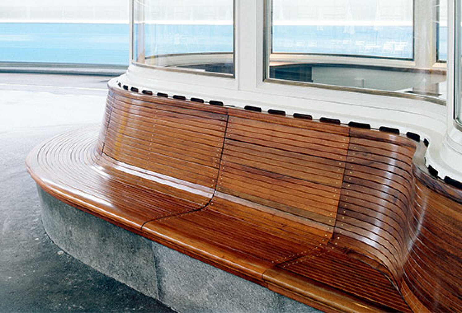 Curved wooden bench with smooth, polished slats, built around a glass-walled structure in a modern indoor space with a concrete base and blue accents in the background.