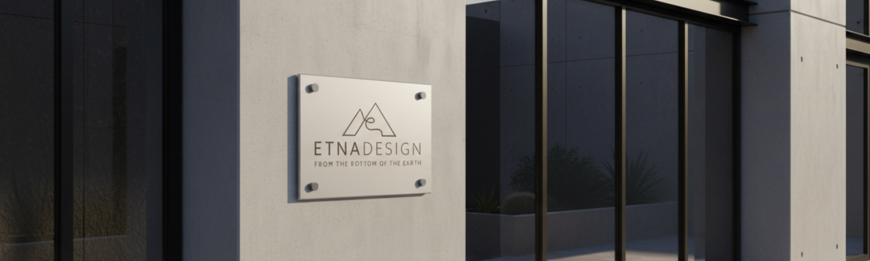 Eine moderne Gebäudefassade mit einem Metallschild mit der Aufschrift ETNADESIGN und dem Slogan FROM THE BOTTOM OF THE EARTH sowie einem minimalistischen Berglogo, die neben großen reflektierenden Glasfenstern angebracht sind.