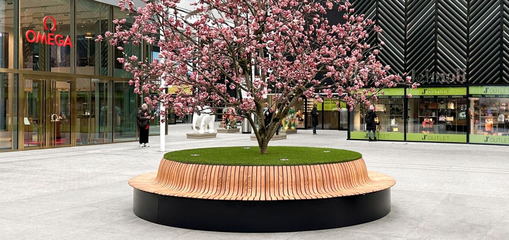 A blossoming pink cherry tree stands at the center of a circular wooden bench surrounded by a modern outdoor shopping area with store signs and glass windows in the background.