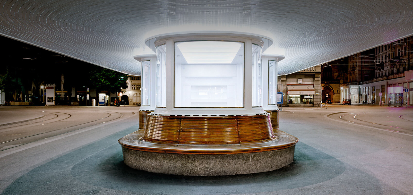 A futuristic, circular, glass and metal pavilion sits empty on a curved street at night, illuminated brightly from within and surrounded by tram tracks and city buildings.