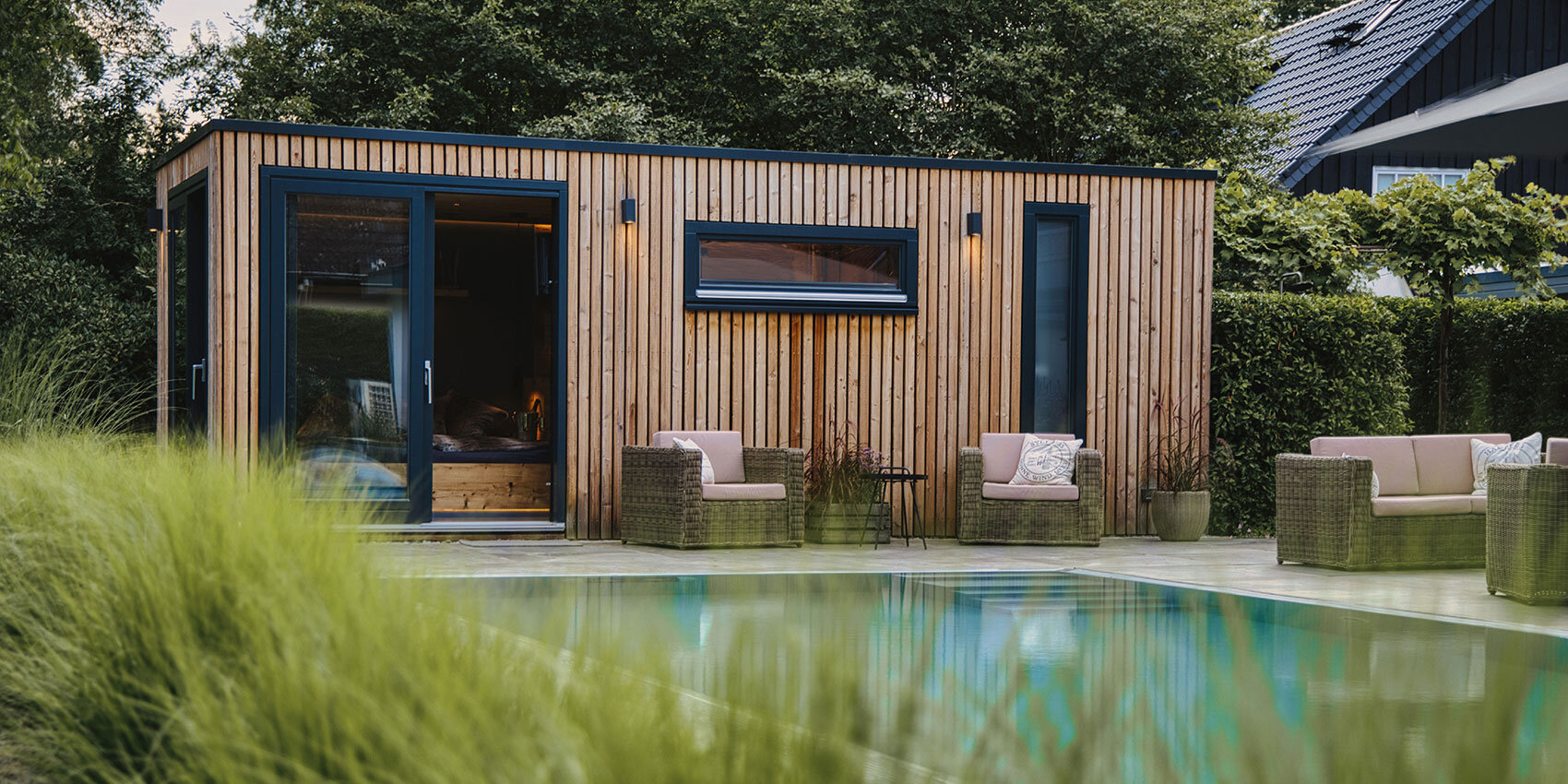 Modern wooden cottage by the pool with large windows, surrounded by wicker sofas and chairs with cushions, lush green bushes and a clear swimming pool in the foreground.