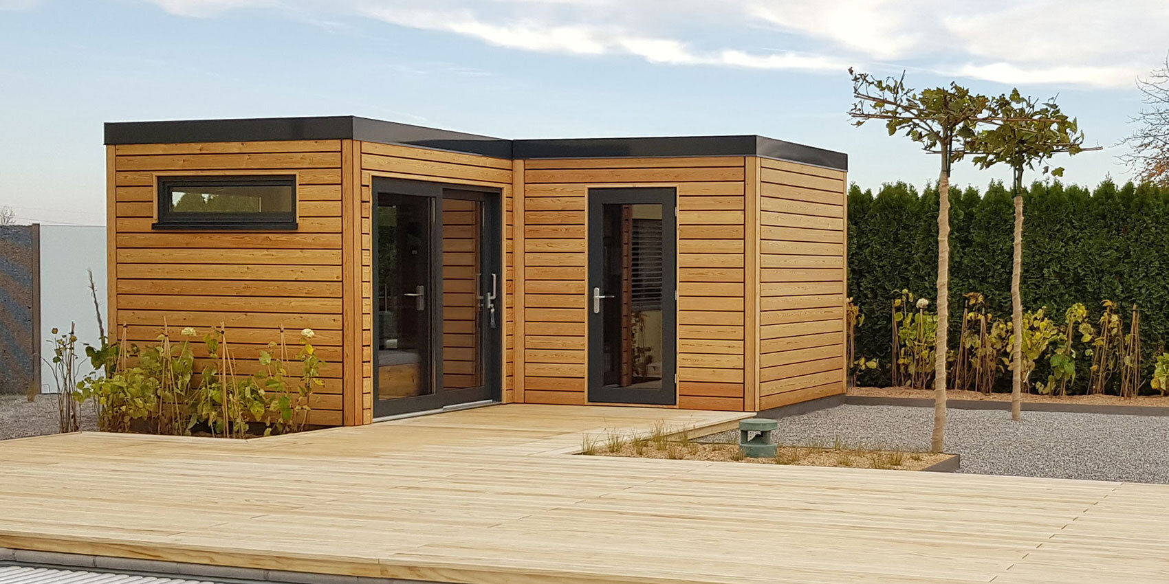 Ein modernes, kleines Holzgebäude mit großen Fenstern und Flachdächern steht auf einem geräumigen Holzdeck, umgeben von Kies, Pflanzen und Bäumen. Eine hohe Hecke säumt den Hintergrund unter einem teilweise bewölkten Himmel.