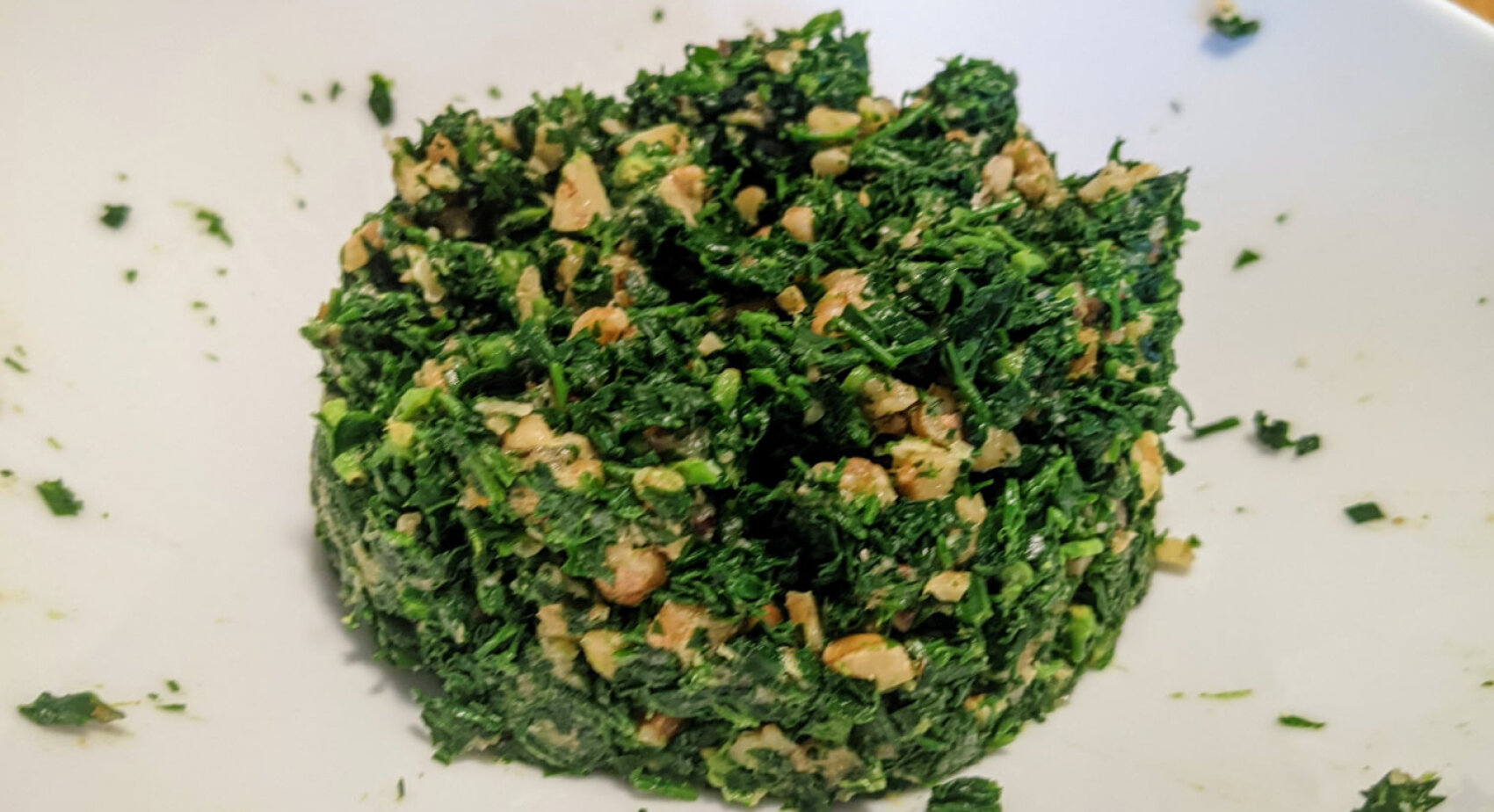 Auf einem weißen Teller befindet sich ein Haufen fein gehacktes, gemischtes Grünzeug und Nüsse. Die Mischung wirkt frisch und leicht feucht, mit sichtbaren Gemüsestücken und knusprigen Stücken.